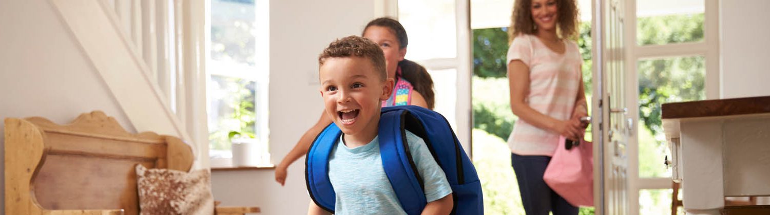 Kids running into house with a backpacks, mom stands in the doorway smiling