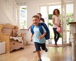 kids running into house with backpacks while mom stands in doorway smiling