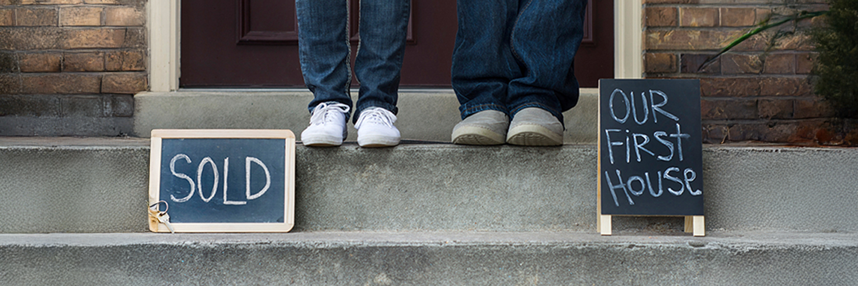 Image of two people's feet on steps with two signs beside them reading "Sold" and "Our first house"